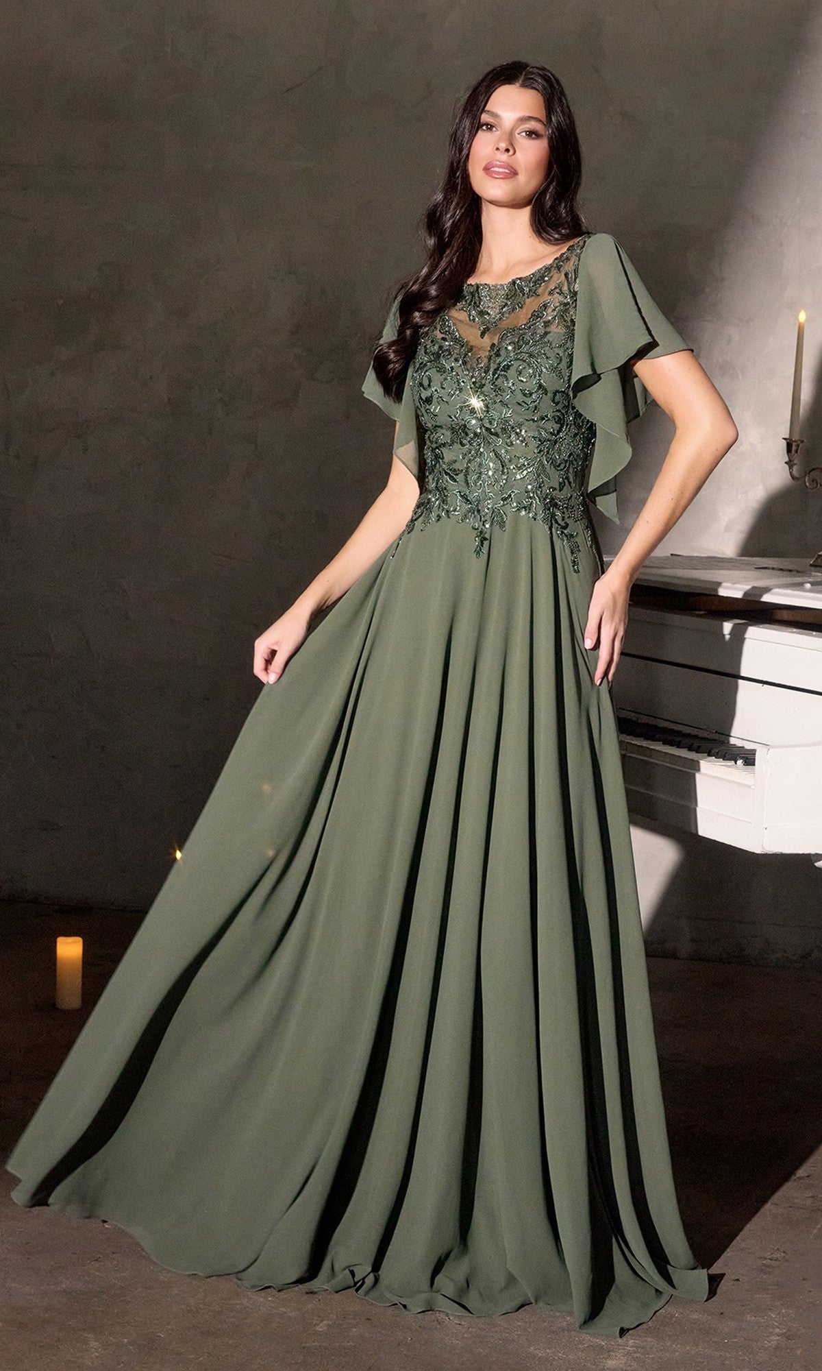 A woman in a long, olive Ladivine CL09 formal dress with lace and flutter sleeves stands indoors by a white piano and candle.
